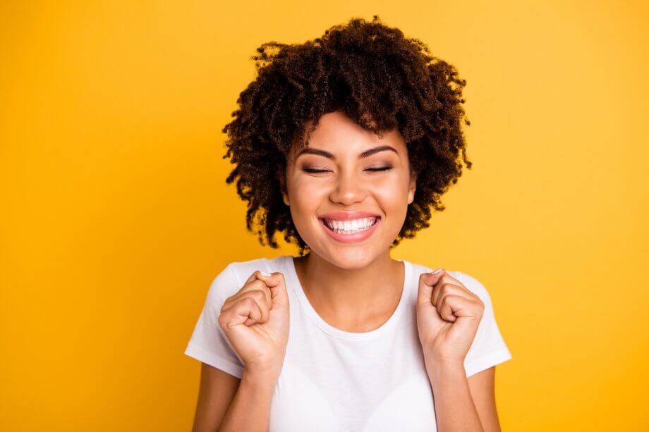 A woman standing in front of a yellow background wearing a white shirt, smiling with her eyes closed.